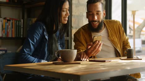 Vorderansicht-Eines-Glücklichen-Jungen-Paares-Gemischter-Abstammung,-Das-Am-Tisch-Im-Café-über-Ein-Mobiltelefon-Diskutiert,-4K