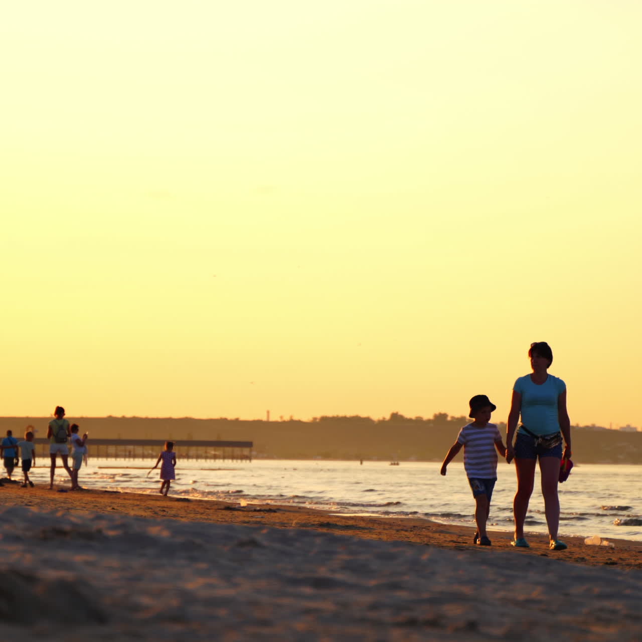 Mother with son are walking near the sea at sunset. Sealine with people in the evening in summer. Enjoyable vacation at the seaside.