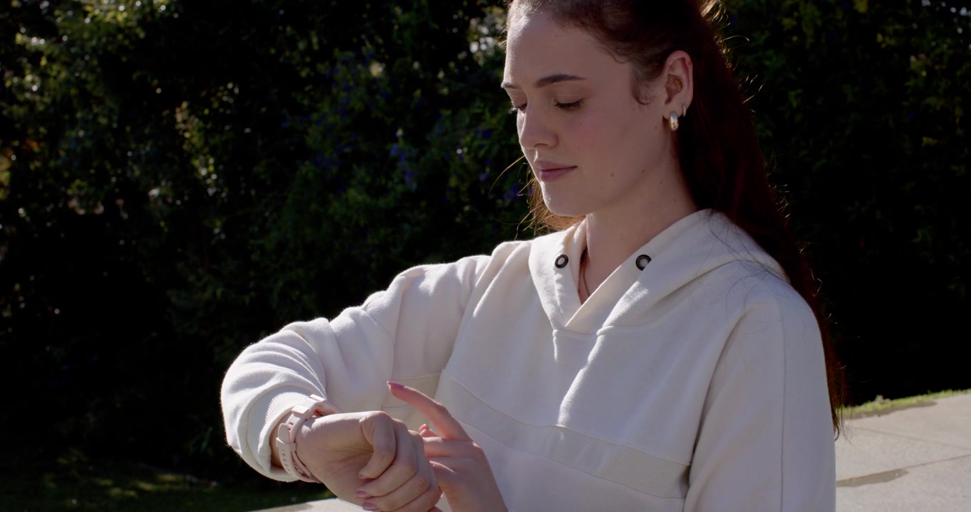 Donna sui vent'anni con felpa con cappuccio color panna, che si ferma all'aperto per toccare lo smartwatch e controllare l'ora