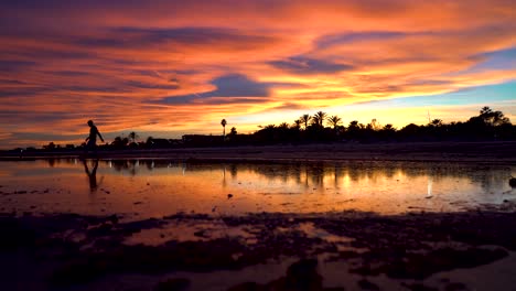 Man-tiptoes-across-a-lake-in-the-center-of-an-incredible-beach-at-sunset,-the-sky-and-the-sun-at-fall-show-amazing-colors-as-he-passes-from-side-to-side