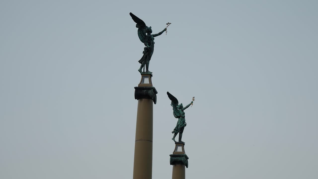 Bronze winged angel statues holding wreaths stand atop tall columns against the evening sky in Prague