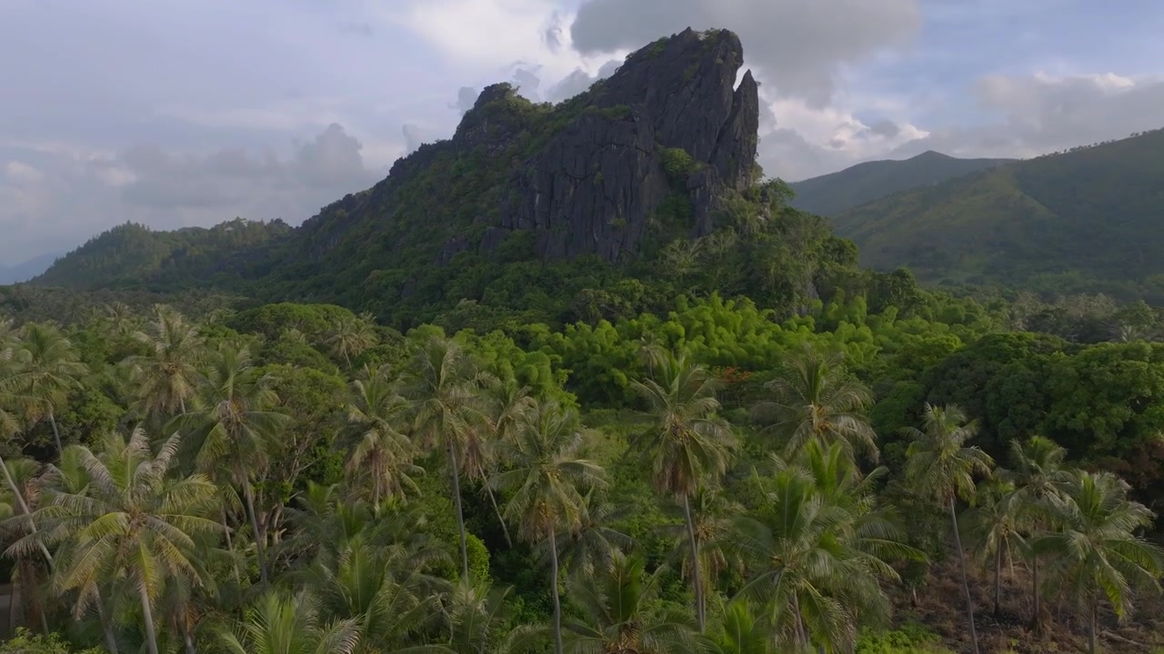 Ripresa aerea molto bassa sopra palme da cocco e vegetazione tropicale, con un'inclinazione lenta verso l'alto per rivelare un'imponente formazione rocciosa contro rigogliosi pendii montuosi.