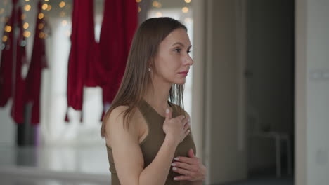 meditation coach sits on yoga mat with hands on chest and stomach, eyes closed in tranquil breath, surrounded by red aerial hammocks in bright studio creating serene mindful practice atmosphere