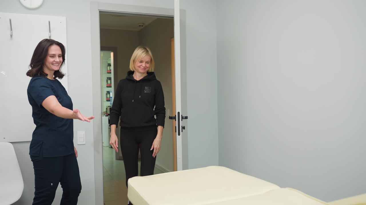 Massage therapist in navy scrubs opens door with warm smile to welcome client into bright white treatment room with modern minimalist decor, soft lighting, and inviting professional atmosphere