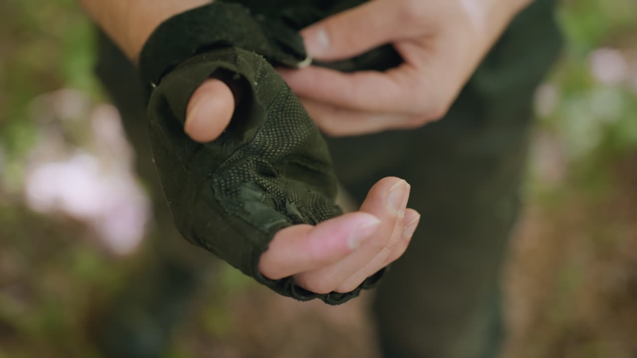 Close up view of warrior palm putting on fingerless glove, veins visible on wrist, blurred forest surroundings, belt and cargo pants visible under dappled sunlight casting textured shadows