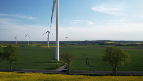 Antena-Hacia-Atrás-Cerca-De-Enormes-Turbinas-Eólicas-Situadas-En-Campos-Agrícolas-Y-Cerca-De-Un-Campo-De-Colza-De-Maduración-Amarilla