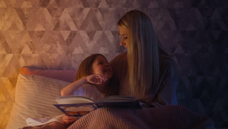 Funny-daughter-holding-book-on-bed-touches-mom-lips-for-kiss