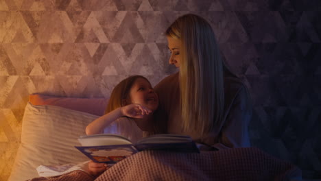 funny daughter holding book on bed touches mom lips for kiss