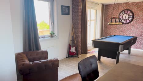 A smooth camera pan reveals a vintage-inspired apartment lounge with a billiard table, exposed brick wall, retro decor, and adjacent modern kitchen in bright daylight
