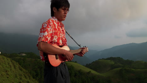 close up shot of an asian man standing on top of a mountain and playing ukulele while singing on a cloudy day