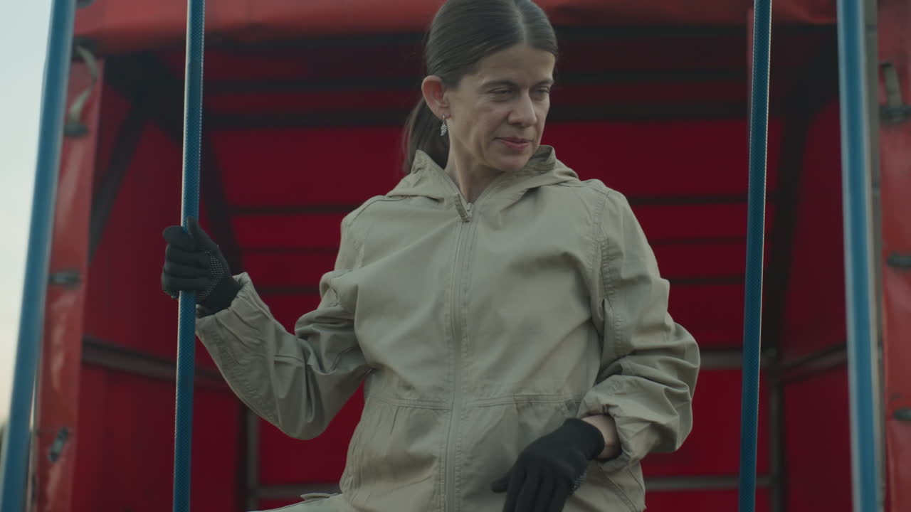 close up of woman inserting blue support poles into wicker balloon basket by red trailer on grassy field under soft sky, focus on careful hand placement and preparation ritual before flight