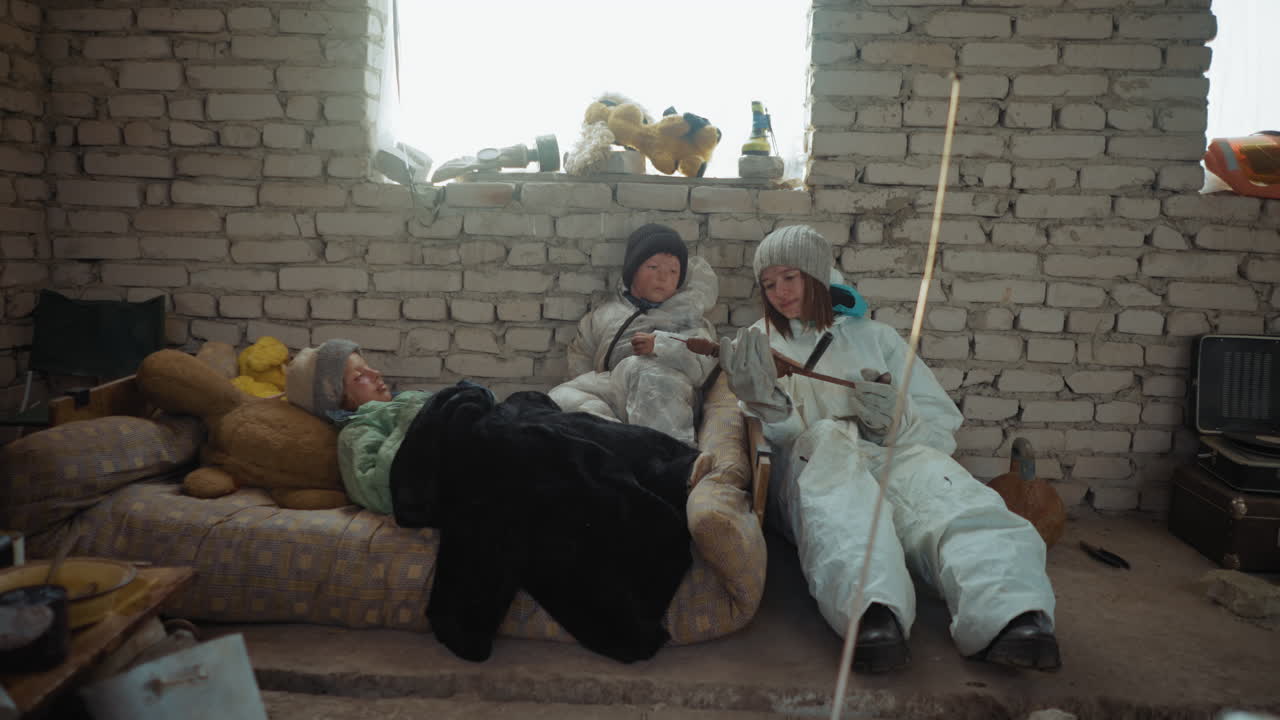 Displaced family huddled inside cold brick shelter with broken window, mother in hazmat suit holding stick while comforting two children dressed in winter clothing lying on mattress