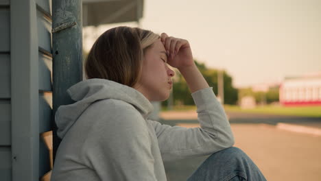 close-up of young lady with hand on head, eyes closed, in thoughtful expression, background is softly blurred, showcasing distant building, trees, and pole