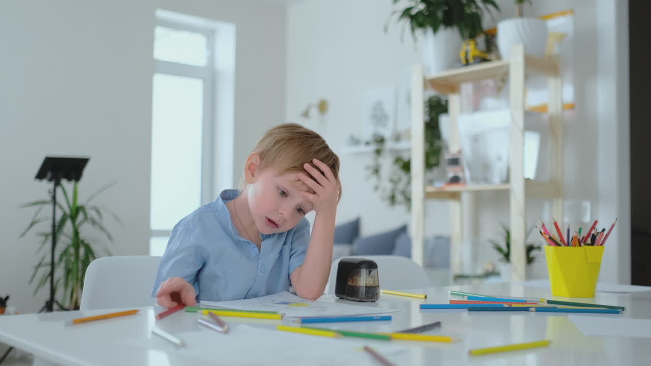 The boy sitting at the table in the living room writes a letter to his father and draws a picture of the family with a pencil