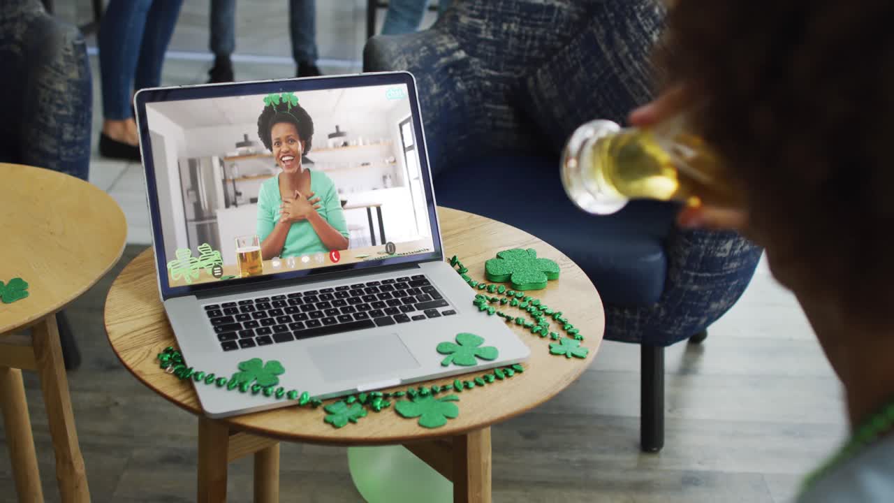 Smiling african american woman with beer wearing clover shape band on video call on laptop