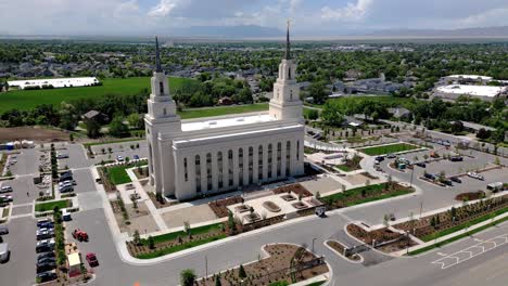 Drone-shot-seeing-full-Layton-LDS-Mormon-Temple-moving-around-to-the-left-reviling-full-city-view-in-middle-of-a-sunny-day-cars-in-parking-lot-and-driving-by-on-road