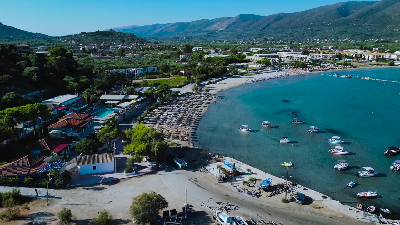 Zoom aéreo para fora revela praia lotada com guarda-sóis de palha, água turquesa cristalina, barcos e hotéis resort com colinas verdes ao fundo