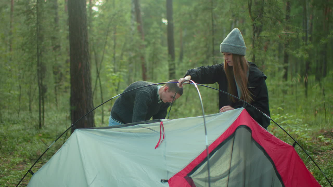 teammates work together setting up camp tent in woodland as woman holds flexible tent pole steady while man adjusts fabric background features trees with soft light filtering through canopy