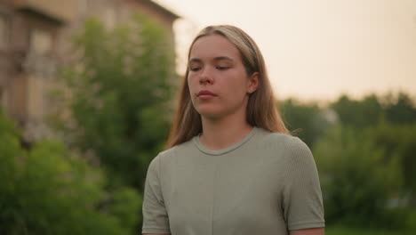 close-up of young woman walking outdoors with a tired expression, dressed casually, against blurred background of greenery, power lines, and distant building