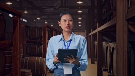 asian female professional worker standing with her tablet in cellar of winery, she observes by looking around before she come to concentrating with her tablet