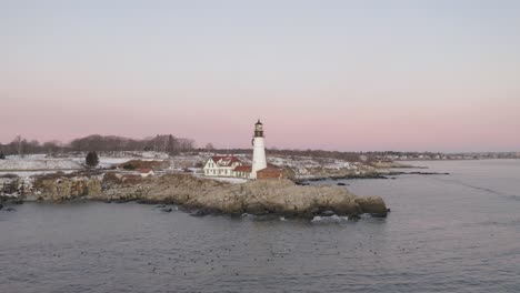 Lighthouse-on-rocky-coastal-outcrop-above-floating-sea-ducks,-winter-sunrise,-SLOW-AERIAL-ORBIT