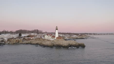 lighthouse on rocky coastal outcrop above floating sea ducks, winter sunrise, slow aerial orbit