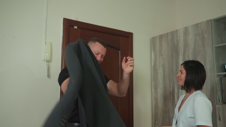clinic entrance therapist helping client remove coat. preparation for microcurrent massage treatment, calm professional atmosphere, soft lighting, wooden door, uniformed woman assists muscular man