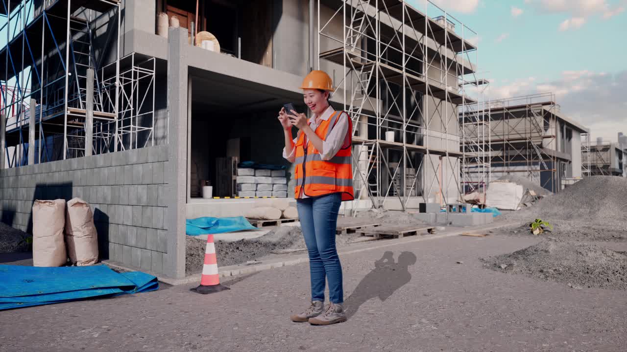 Full Body Side View Of Asian Female Engineer With Safety Helmet Using Smartphone While Standing At Construction Site in Progress to New House
