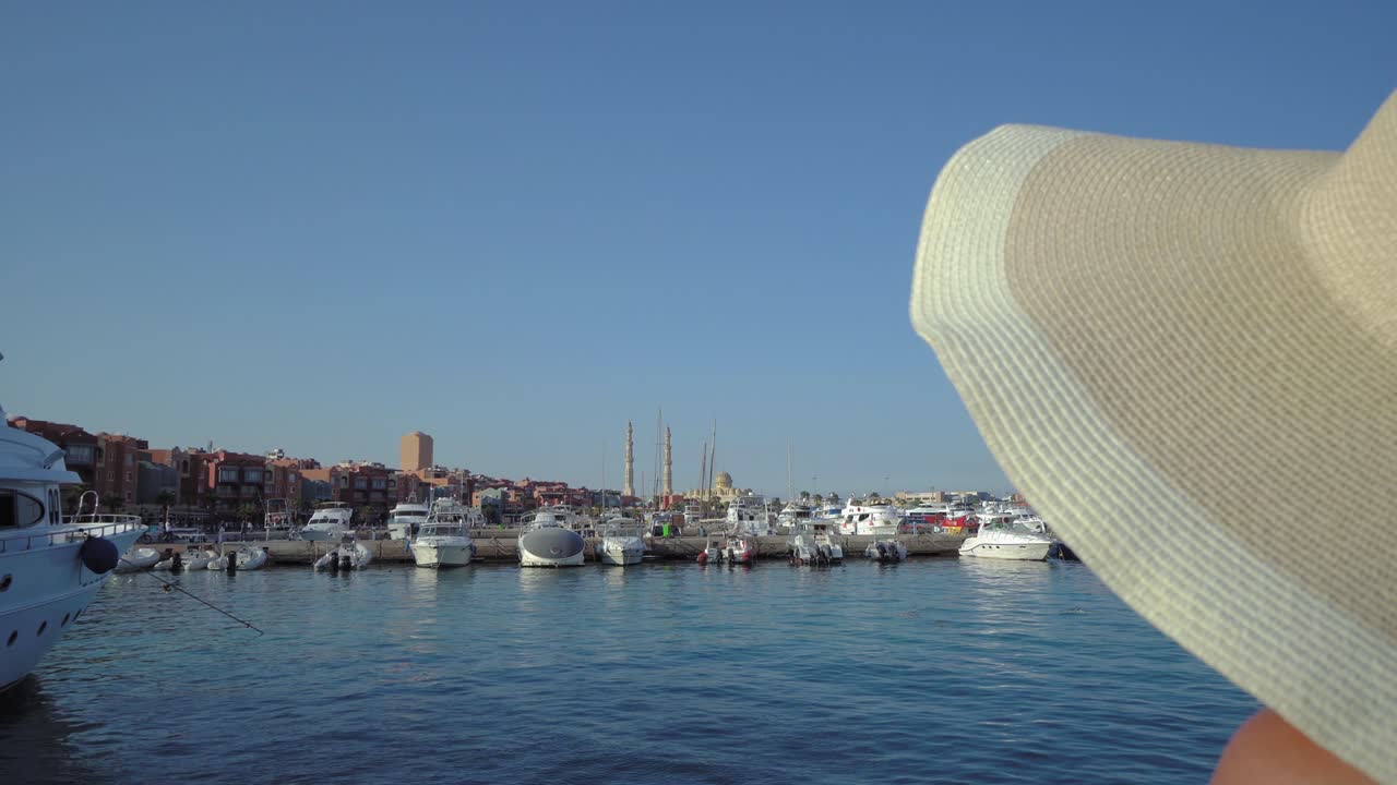 모자를 입은 여성이 이집트의 허르가다 마리나 (hurghada marina) 의 전망을 즐기고 있으며, 부두에 있는 배와 요트, 뒷면에는 무슬림 모스크 엘 미나 (el mina) 가 있다.