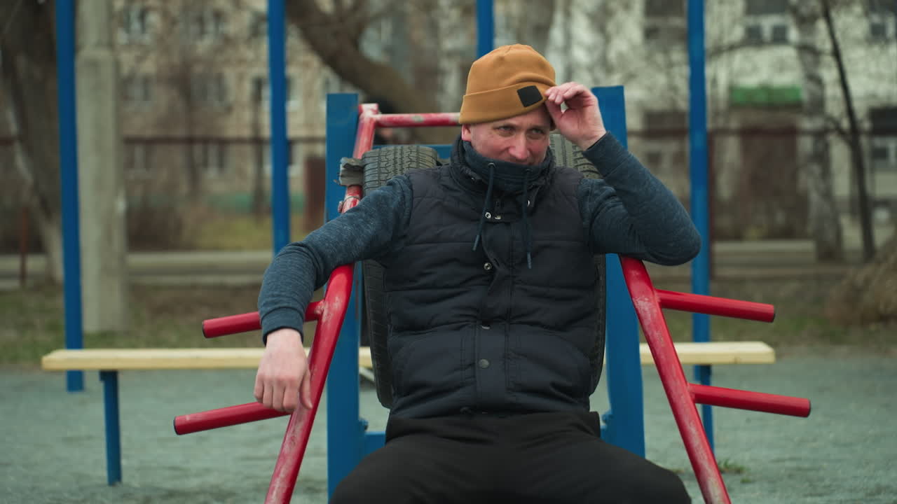 A coach seated on workout equipment, adjusting his beanie, looks tired, in the background, a boy dressed in black plays football