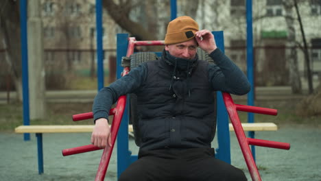 a coach seated on workout equipment, adjusting his beanie, looks tired, in the background, a boy dressed in black plays football