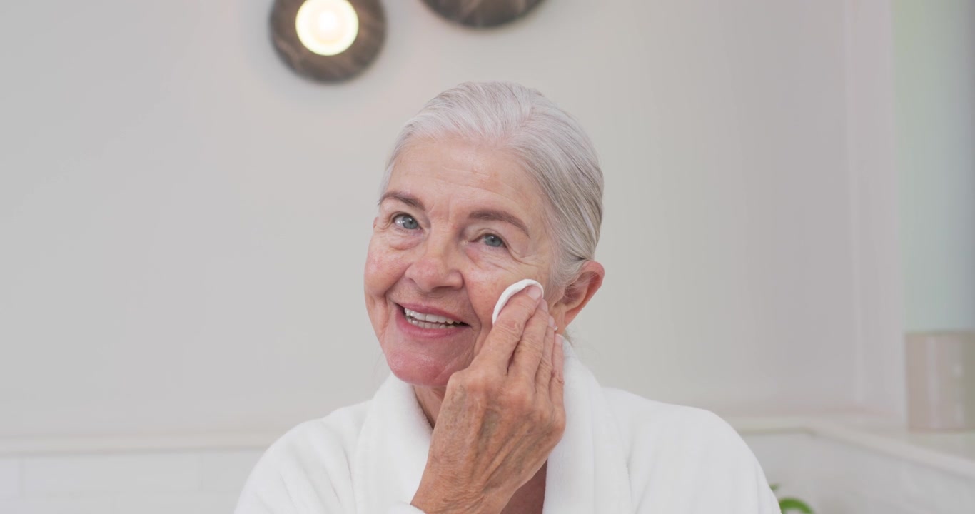 Senior female wearing bathrobe starting skincare at bathroom vanity, wiping face with cotton pad