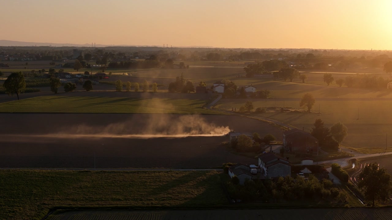Vista aerea laterale con drone della lavorazione del terreno agricolo che genera grandi nuvole di polvere su campi aridi nella Pianura Padana, vicino a Cremona, Lombardia, Italia settentrionale, durante la luce del tramonto primaverile, paesaggi rurali.