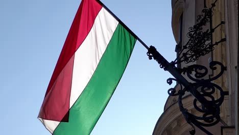 red, white and green stripes of the hungarian flag flutter quietly from a gustatory pole on a budapest facade on a sunny day in the wind