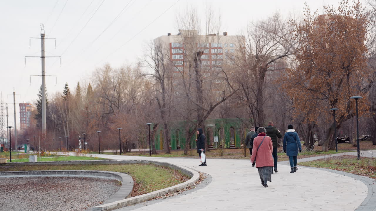 People stroll along winding park walkway lined with bare trees and autumn leaves, passing lamp posts, benches and power lines near apartment buildings, under overcast sky during quiet late afternoon