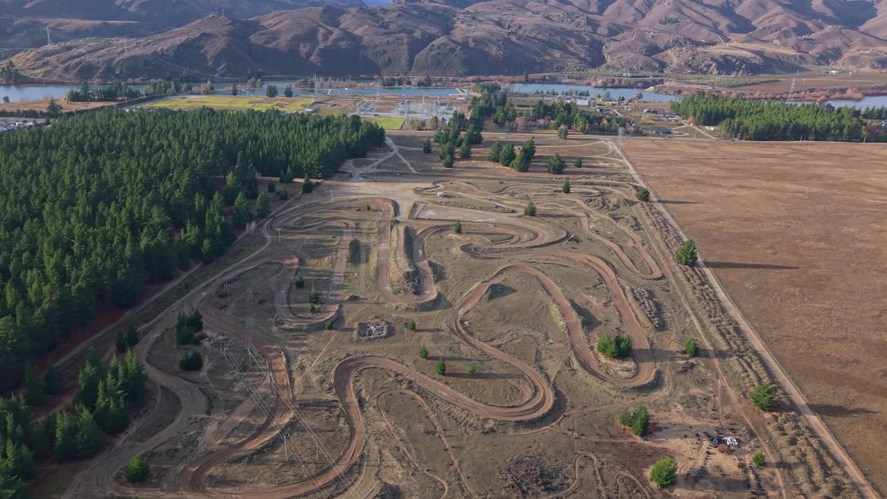 Aerial footage of a motocross track near Cromwell, New Zealand, featuring winding dirt circuits, pine forests, farmland, Lake Dunstan, and dramatic Central Otago mountains.