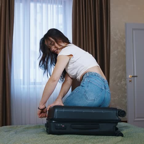 Woman closes her full suitcase in the hotel. Happy young woman in jeans and white t-shirt sits on a travel bag to close it before leaving the hotel room