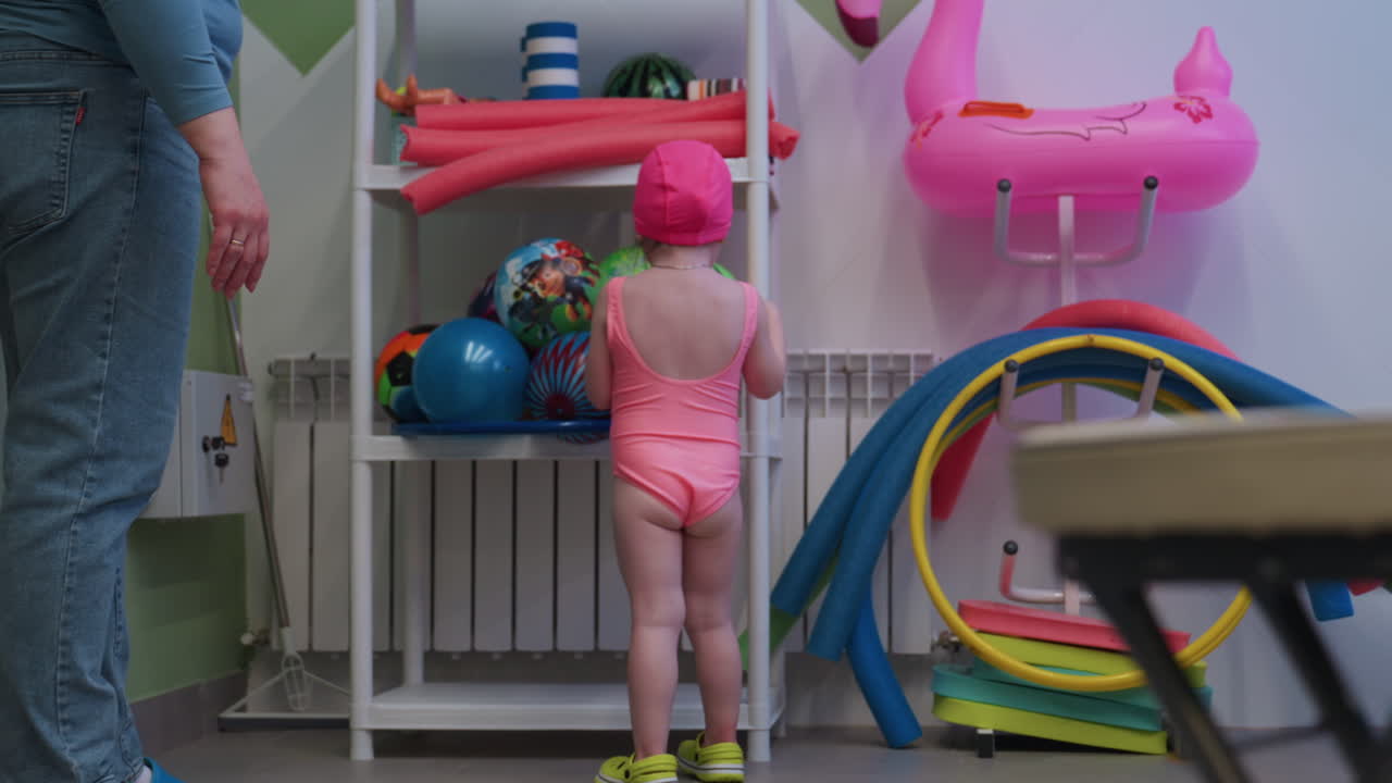 Kid following mother toward spot where air balls packed, tiny steps, hand reaching, curious gaze, poolside light, playful anticipation, side view, family outing, guidance toward stacked toys
