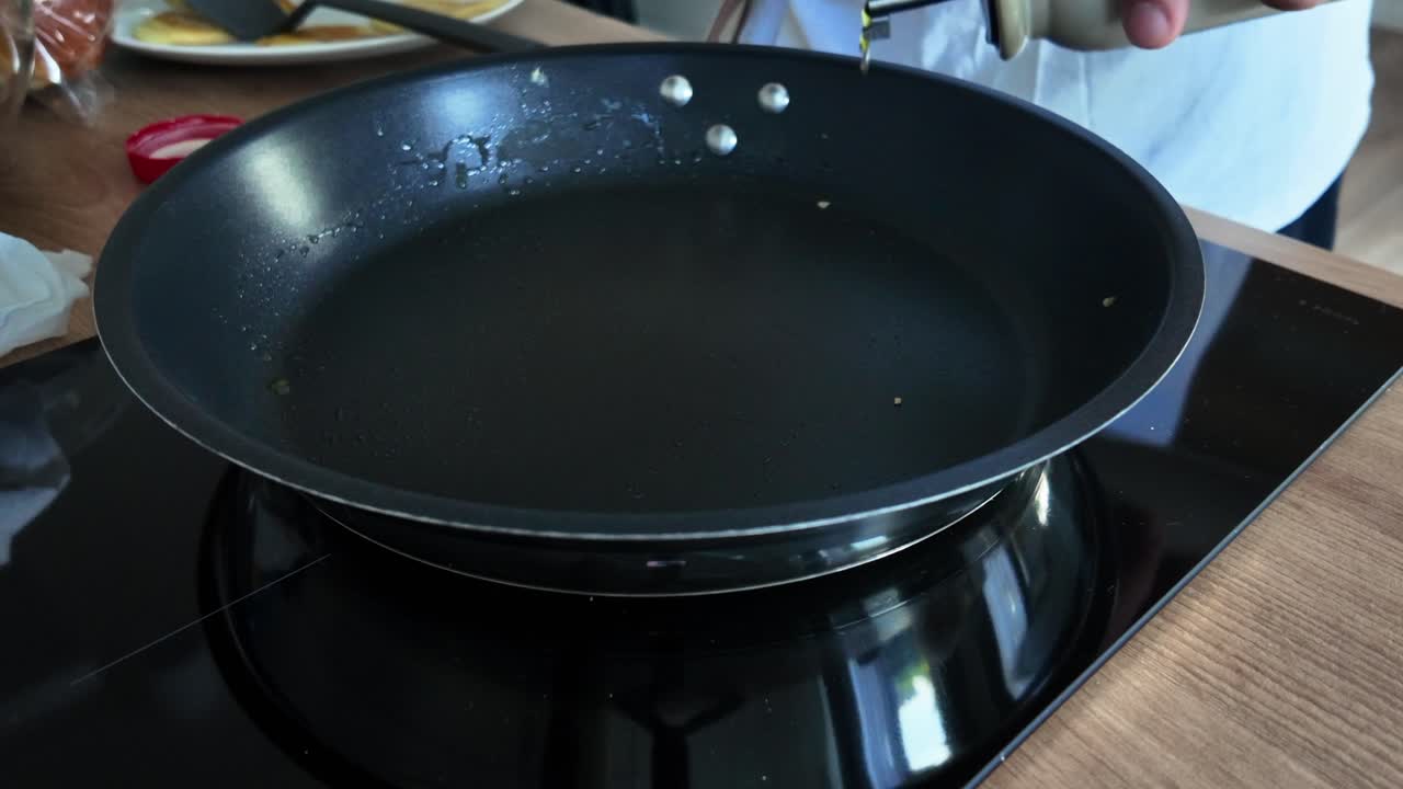 Close-up of oil being poured into a black nonstick frying pan on a modern induction stovetop in a home kitchen, capturing the beginning of a cooking process