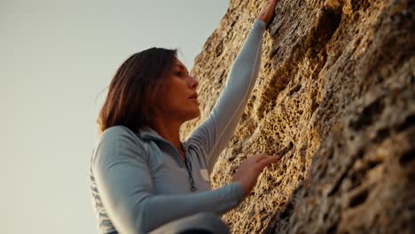 Shot-close-up,-shot-from-below,-a-blonde-girl-in-a-blue-jacket-climbs-a-steep-rock-on-which-the-rays-of-the-sun-fall.-blonde-girl-climber-climbs-up-a-steep-rocky-rock-and-looks-for-where-to-put-her-fingers-for-a-comfortable-spiral