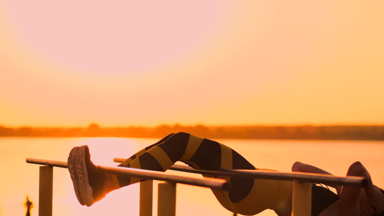 복부 근육 운동: 여름에 호수에 대한 해가 지는 동안 운동하는 젊은 미녀, 바에서 느린 동작으로 복부 운동을 하는 스포츠 옷을 입은 여성