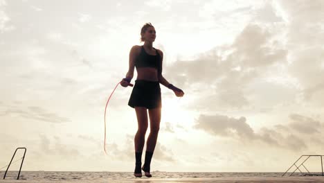 athletic woman in shorts and sport bra working out on the jump rope against the son by the beach in slow motion. girl jumping on