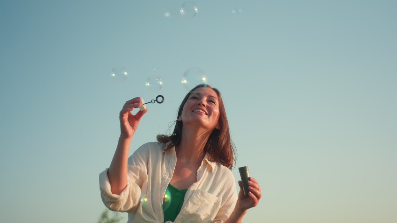 Front view of lady joyfully blowing bubbles under clear blue sky with soft focus tree in background, sunlight illuminating her relaxed expression while bubbles rise gently into air