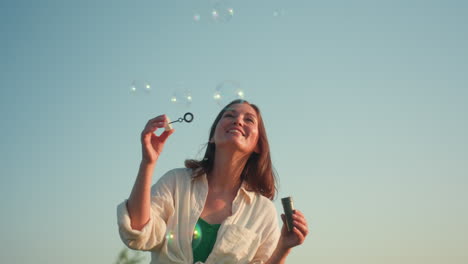 front view of lady joyfully blowing bubbles under clear blue sky with soft focus tree in background, sunlight illuminating her relaxed expression while bubbles rise gently into air