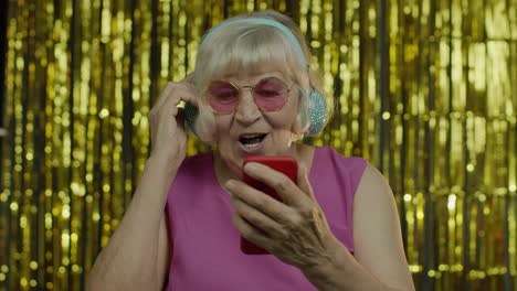 stylish happy senior old woman holding cell phone, listening to music in headphones and dancing