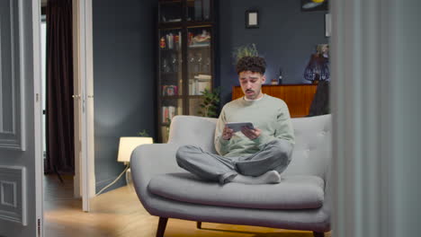 young man sitting on sofa with crossed legs and watching something on smartphone