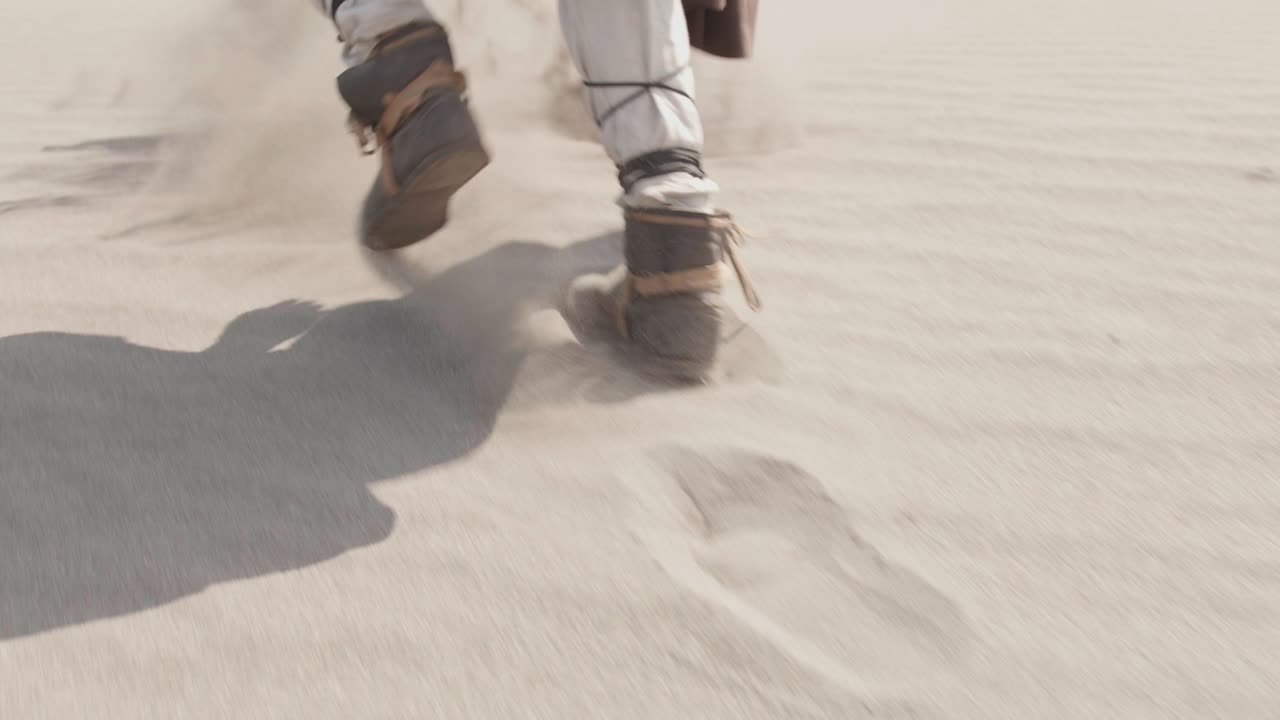 Cinematic slow motion shot following the feet of a robed character walking through a vast, sandy desert. A concept of a long, difficult journey, adventure, or exploration