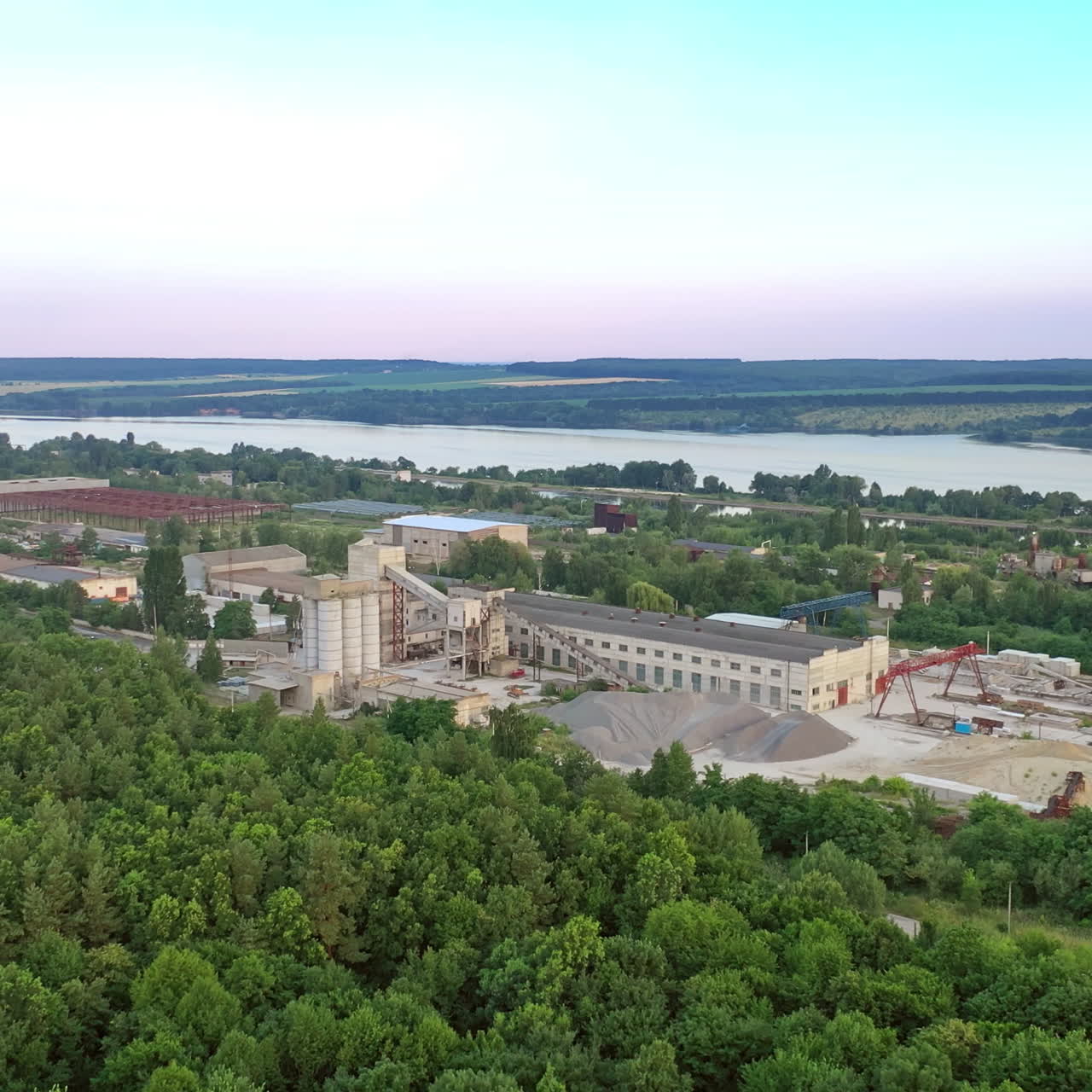 Flying over green trees to industrial territory in summer. Industrial factory near the river on the natural background.