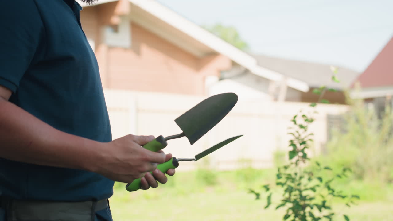 Side view adult gardener tosses hand shovel, tool drops to ground, then he picks it up and knocks dirt by striking against another shovel beside waist apron pouch, backyard fence