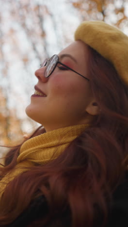dama sonriente con una boina amarilla y abrigo negro ajustando una hoja de otoño vibrante, mirando hacia arriba con una expresión alegre en un bosque iluminado por el sol